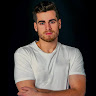 Portrait of a man with short dark hair and beard wearing a white t-shirt with arms crossed against a black background.